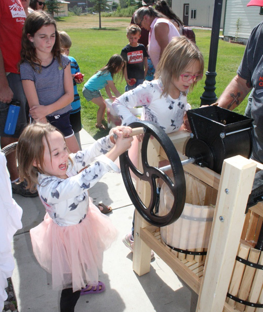 color image a little girl in a white top and pink tutu cranking the handle on a cider press with a huge smile on her face