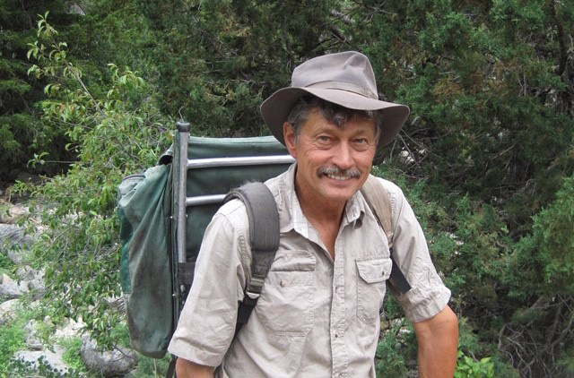 color image of a man in a tan shirt and hat wearing a green backpack standing in a forest