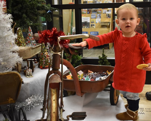 color image of a toddler dressed in red standing next to a historic tricycle in front of a Christmas Tree